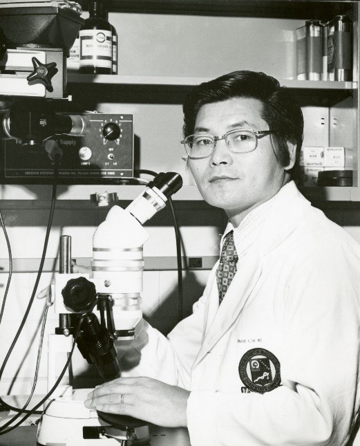 Black and white photograph of scientist Dr. David J. Lim in a lab coat using a microscope in his laboratory at Ohio State.