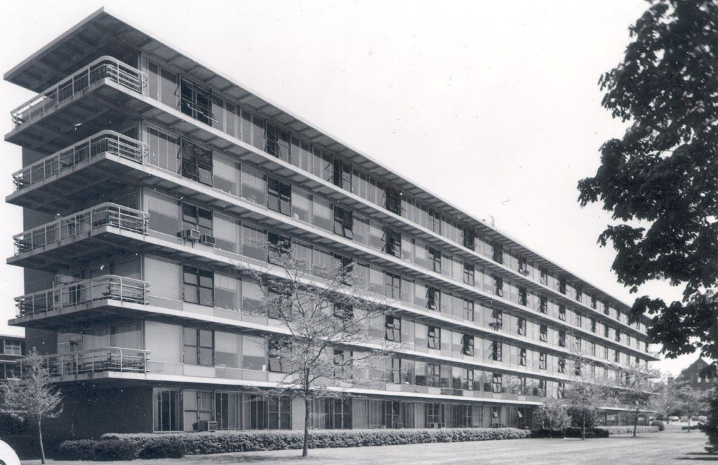 Black and white photograph of a long, rectangular modernist building with five floors and continuous balconies along each level. The building is surrounded by trees and shrubs.